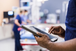 Doctor reviews a tablet in the foreground while other doctors and nurses stand over a medical bed in the background