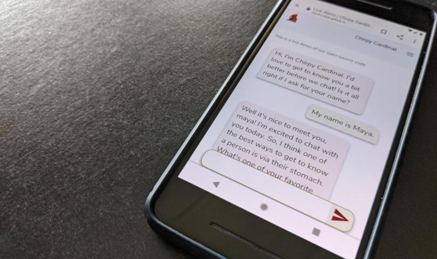 A phone on a desk displays a text chat with Chirpy, a Stanford chatbot.