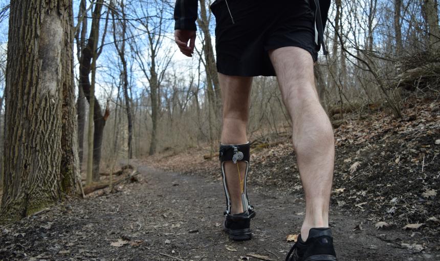 A man walks in a forest with an ankle exoskeleton attached to his left leg. 