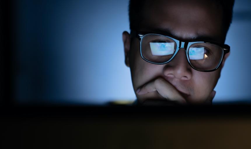 A man wearing glasses stares at his computer screen in a dark room. 