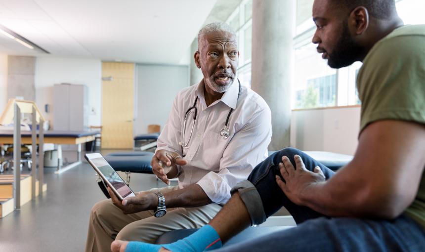 Serious male orthopedic doctor uses a digital tablet to show an injured male patient a video about appropriate exercises to strengthen the patient's food and ankle. The patient's food and ankle are wrapped with kinesiology tape.
