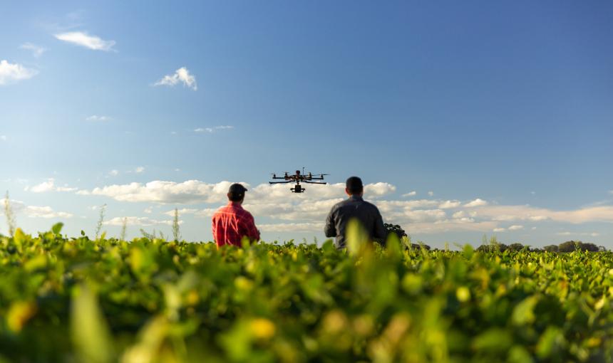 Two farmers stand in a field with a drone over their heads.