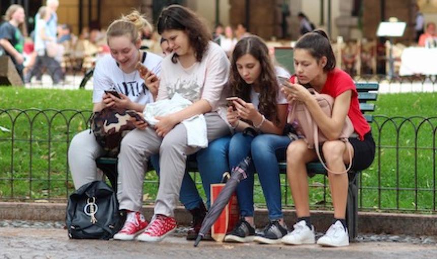 Young women line a park bench, each engrossed in her smartphone.