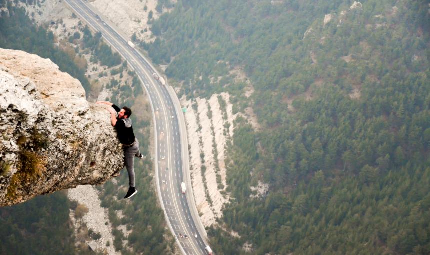 Man hangs off a mountain cliff, dangling dangerously with a road far below