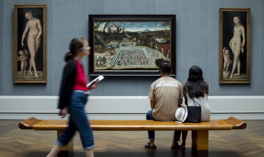 A couple sit on a bench in a museum looking at a work of art.
