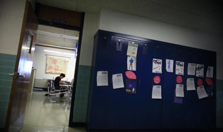 A student studies at a desk inside a classroom next to blue lockers. 
