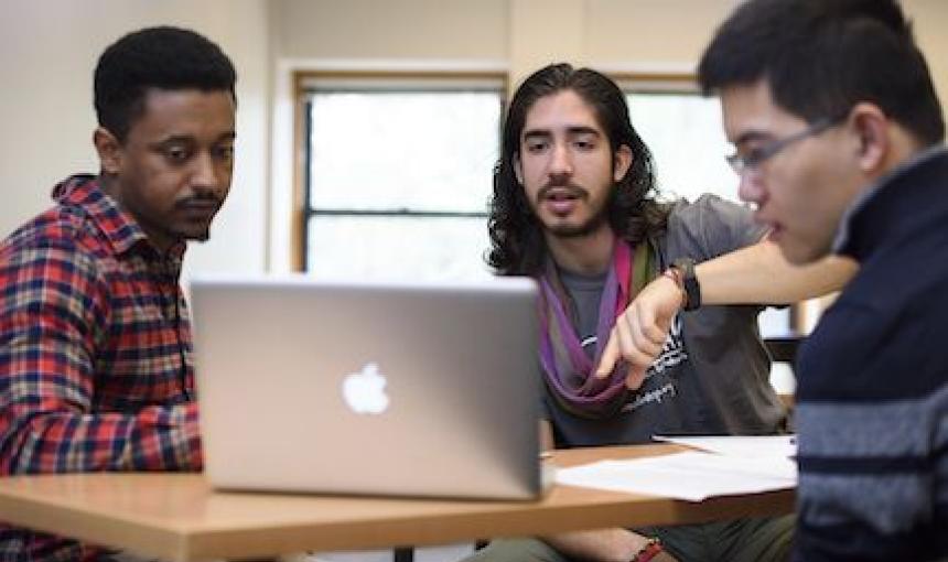 Three student researchers discuss their work while looking at a computer and sitting at a table. 