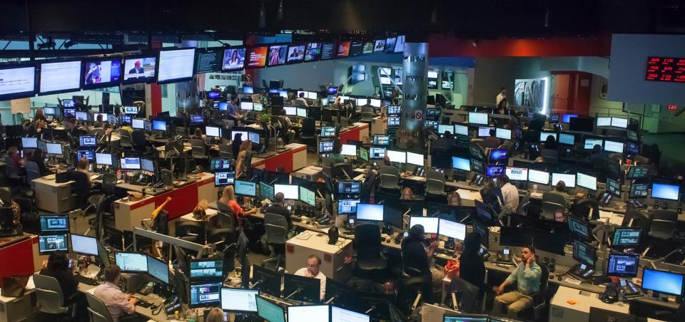 a shot of a busy newsroom filled with desks, computer screens, and people.
