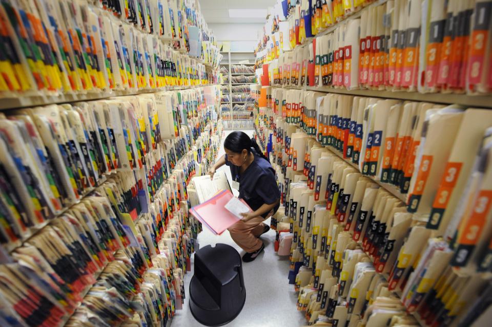 A woman sits between two shelves overflowing with medical records. 
