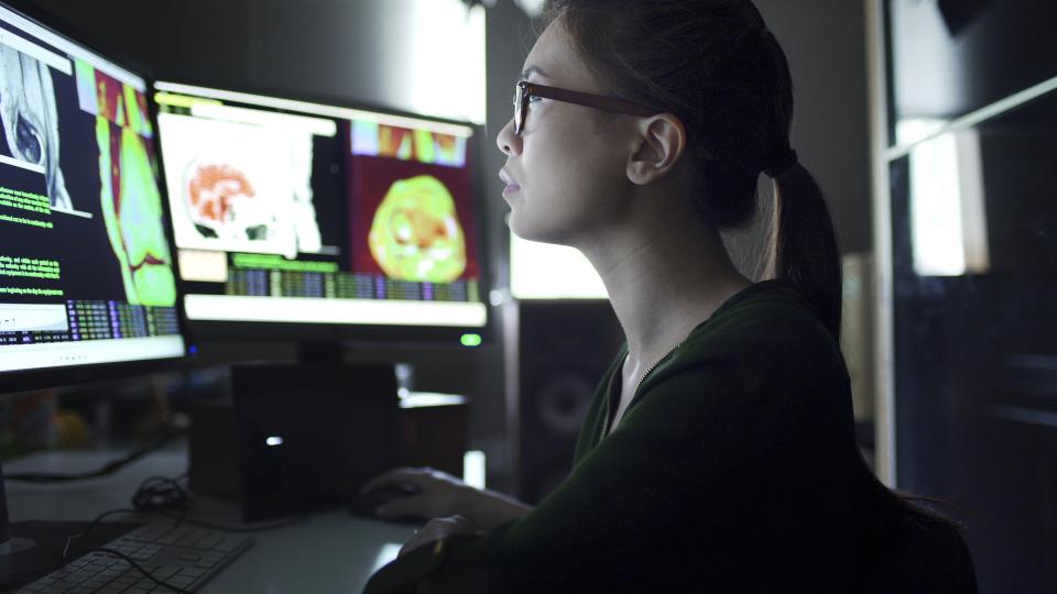 A young woman working at a desk with multiple monitors studies data & information on the screens which consists of MRI & CAT scans.