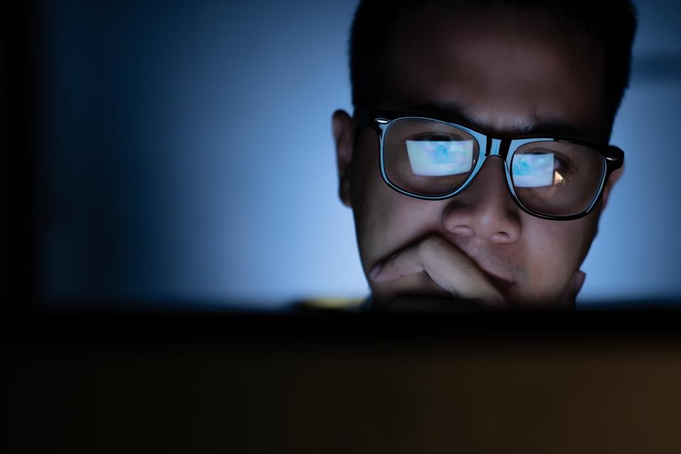 A man wearing glasses stares at his computer screen in a dark room. 