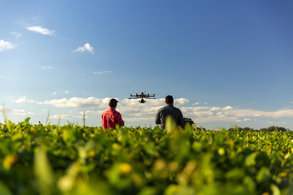 Two farmers stand in a field with a drone flying above them