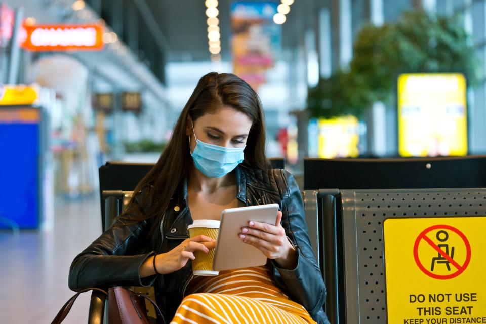 young woman traveling by plane during COVID 19, wearing N95 face mask, using a digital tablet in airport waiting area.