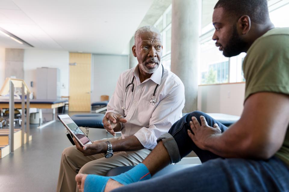 Serious male orthopedic doctor uses a digital tablet to show an injured male patient a video about appropriate exercises to strengthen the patient's food and ankle. The patient's food and ankle are wrapped with kinesiology tape.