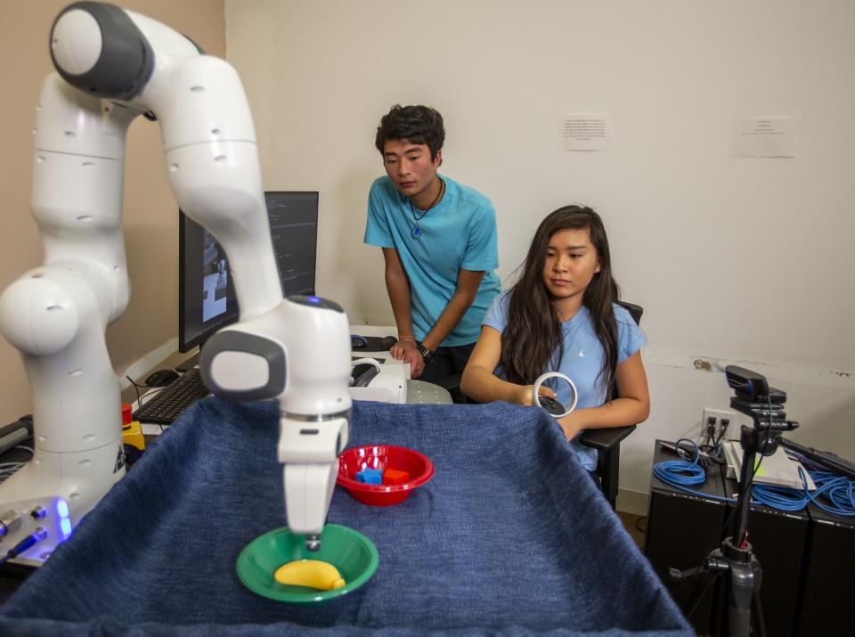 Two Stanford students sit behind a large robotic arm and practice moving blocks with it.